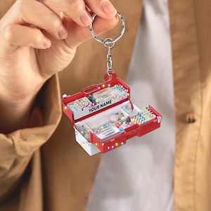 May include: A red and white keychain shaped like an open medical kit, with the text "YOUR NAME" on the front. The kit is filled with miniature medical supplies and is decorated with colorful lights. The keychain is held by a silver ring.
