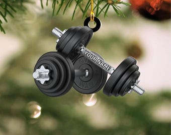 May include: Black dumbbell Christmas ornament with the text "YOUR NAME" on the bar. The ornament is hanging from a gold string against a blurred background of a Christmas tree and ornaments. The dumbbell has weight plates.