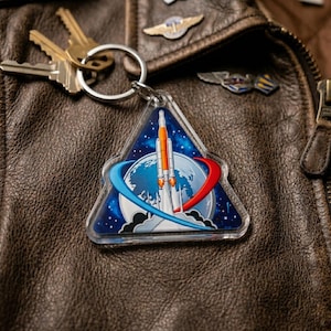 May include: A triangular keychain featuring a rocket launch design with a blue, white, and orange color scheme. The keychain is attached to a silver keyring with gold keys. The background is a brown leather jacket.