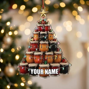 May include: A Christmas tree-shaped hanging ornament constructed from stacked conga drums in various shades of red, black, and natural wood. The ornament features a gold star at the top, string lights, and the text YOUR NAME printed at the bottom.