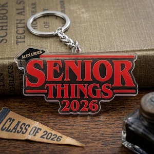 May include: A clear acrylic keychain with the words "SENIOR THINGS 2026" in red lettering. The keychain has a silver keyring and chain. A small pennant that says "CLASS OF 2026" is also visible.