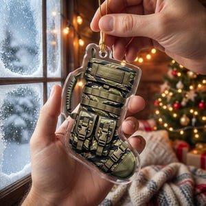 May include: A clear acrylic Christmas stocking ornament featuring a tactical design with a tiger stripe camouflage pattern, integrated webbing straps, a side handle, and two front pockets with plastic buckle closures, suspended by a gold cord.