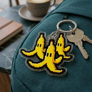 May include: A clear acrylic keychain featuring a pixelated design of three yellow bananas with black accents. The keychain is attached to a silver keyring with keys and is resting on a teal backpack.