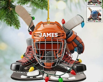 May include: A hockey-themed Christmas ornament featuring an orange helmet with the name "JAMES", skates, a hockey stick, and gloves. The ornament is decorated with a string of colorful Christmas lights and is hanging from a gold string. The text "PERSONALIZED DESIGN" is at the top.