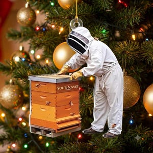 May include: A flat, hanging ornament featuring a beekeeper in a white protective suit and veil, standing beside a stacked wooden beehive. The hive has the text YOUR NAME printed on the front, with small bee illustrations on the orange panels.