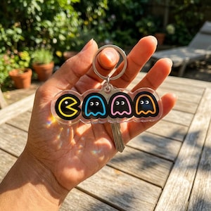 May include: A clear acrylic keychain featuring four colorful Pac-Man ghost characters. The keychain has a silver metal ring and a key attached. The ghosts are yellow, blue, pink, and orange with black outlines.