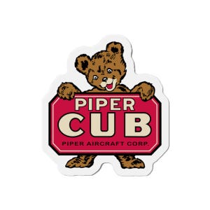 Könnte beinhalten: Ein Aufkleber mit einem braunen Bären, der ein rotes Schild mit den weißen Worten "PIPER CUB" hält. Der Bär hat einen lächelnden Ausdruck und der Text "PIPER AIRCRAFT CORP." steht unten auf dem Schild.
