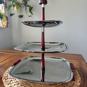 May include: A three-tiered serving tray with a polished silver finish and dark red wooden accents. The tray has a central support and three square-shaped tiers. The tray is sitting on a woven placemat.