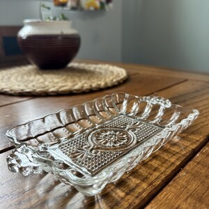 Peut inclure: Un plat de service rectangulaire en verre transparent avec un motif floral décoratif au centre. Le plat a un bord festonné et des poignées à chaque extrémité. Le plat est posé sur une table en bois.