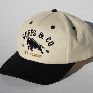 Puede incluir: Gorra de béisbol color crema con visera negra. La gorra presenta el texto "BUFFS & CO." y un gráfico de un bisonte. También se muestra la frase "WE COMIN'". La gorra tiene un botón negro en la parte superior.