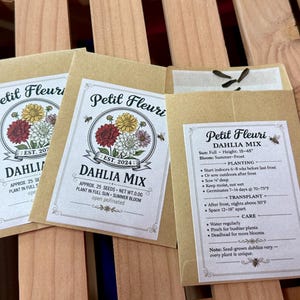 May include: Three seed packets with the text "Petit Fleuri DAHLIA MIX" and a floral illustration. The packets are tan with white labels and feature planting instructions and seed details. The packets are on a wooden surface.