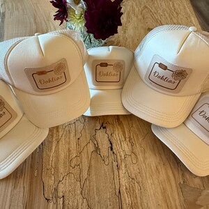 Peut inclure: Plusieurs casquettes de camionneur crème avec un dos en filet et une visière incurvée. Chaque casquette présente un écusson rectangulaire avec le mot "Dahlias" et une illustration florale. Les casquettes sont disposées sur une surface en bois.