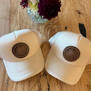 May include: Two cream-colored trucker hats with a brown floral design patch. The hats have a mesh back and a curved brim. The hats are displayed on a wooden surface, with a vase of flowers in the background. The hats are a stylish accessory.