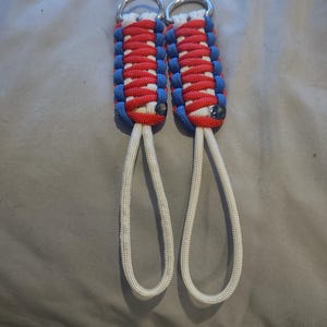 May include: Two handmade keychains with a patriotic color scheme. Each keychain features a woven design in red, white, and blue paracord, with a silver metal ring at the top and a white loop at the bottom.