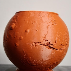 May include: A spherical, orange-brown decorative vase with a textured surface, resembling the planet Mars. The vase has a wide opening and sits on a circular base. The surface features raised bumps and lines, mimicking the planet's terrain.
