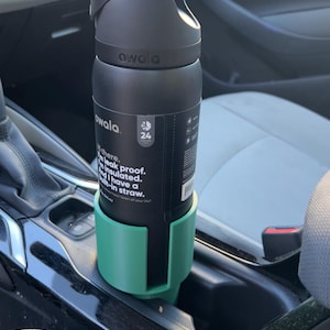 May include: A black Owala water bottle with a handle is in a green car cup holder. The bottle has the brand name printed on it and features a label with the text "leak proof" and "insulated". The bottle is in a car.