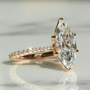 May include: A rose gold engagement ring featuring a large, marquise-cut diamond as the centerpiece. The band is adorned with smaller, sparkling diamonds. The ring is set against a soft, reflective background, highlighting its brilliance.
