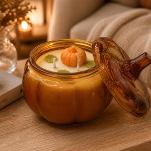 May include: A pumpkin-shaped candle in a glass jar with a matching lid. The candle has a pumpkin and leaf-shaped wax decorations. The jar and lid are amber-colored, and the candle wax is a light orange color. The candle is on a wooden surface.