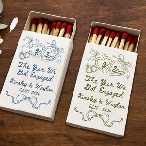 May include: Two white rectangular matchboxes filled with wooden matches featuring red tips. Each box displays blue or green ink illustrations of two hearts, wedding rings, and a bow, with the text The Year We Got Engaged, Kinsley & Waylon, EST. 2026.
