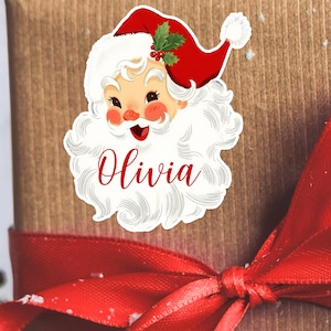 May include: A Santa Claus sticker with a red hat, white beard, and the name "Olivia" in red script. The sticker is placed on a wrapped gift with a red ribbon.