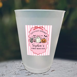 May include: A frosted plastic cup featuring a pink and white striped label with a scalloped border. The label displays a ghost, a pumpkin, a mummy cat, and bats, with the text: Let's Get Spooky, Sophia's First Boo-Day, and various candy icons.