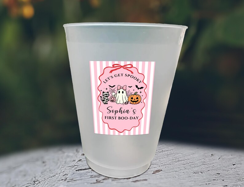 May include: A frosted plastic cup featuring a pink and white striped label with a scalloped border. The label displays a ghost, a pumpkin, a mummy cat, and bats, with the text: Let's Get Spooky, Sophia's First Boo-Day, and various candy icons.