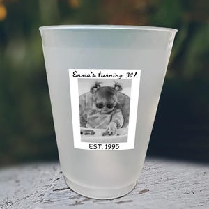 May include: A frosted plastic cup with a black and white photo of a baby wearing sunglasses. The photo is labeled "Emma's turning 30!" and "EST. 1995". The cup is likely for a birthday celebration.