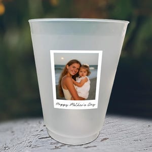 May include: A frosted plastic cup with a square photo of a woman and child on the front. The photo is bordered in white and has the words "Happy Mother's Day" printed below the image. The cup is sitting on a white surface.