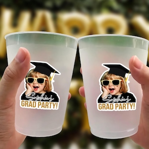 May include: Two frosted plastic cups, each adorned with a sticker featuring a child in sunglasses and a graduation cap. The stickers read "Sophia's GRAD PARTY!" in gold and black. The cups are held up in front of a blurred background.