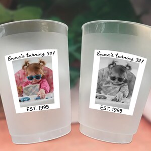 May include: Two frosted plastic cups with a photo label. The label reads "Emma's turning 30!" and "EST. 1995" with a photo of a young child wearing sunglasses. The cups are likely for a birthday celebration.
