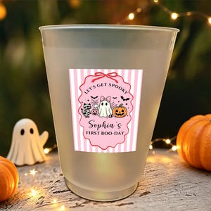May include: A frosted plastic cup featuring a pink and white striped label with a scalloped border. The label displays a ghost, a mummy cat, and a pumpkin, with the text "LET'S GET SPOOKY" and "Sophia's FIRST BOO-DAY" printed in black.