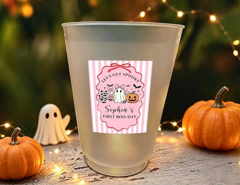 May include: A frosted plastic cup featuring a pink and white striped label with a scalloped border. The label displays a ghost, a mummy cat, and a pumpkin, with the text "LET'S GET SPOOKY" and "Sophia's FIRST BOO-DAY" printed in black.