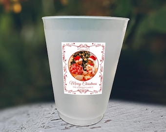 May include: A frosted plastic cup with a rectangular sticker featuring a Christmas photo. The sticker has a red border with the words "Merry Christmas" and "The Harrison Family". The photo shows two people wearing Santa hats.