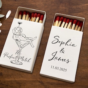 May include: Two white matchboxes with red-tipped matches. One box features a dog illustration in a martini glass with the text "The Perfect Match." The other box has the names "Sophia & James" and the date "11.03.2025" in black script.