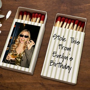 May include: Two matchboxes with red-tipped matches. One box features a photo of a woman wearing sunglasses and holding a drink. The other box has the text "Stole This From Evelyn's Birthday" in black script on a striped background.