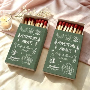 May include: Two rectangular matchboxes with dark green covers featuring white line art of camping gear, trees, and the text "ADVENTURE AWAITS", "Emily & Daniel", and "11.14.2027". The boxes are open, revealing wooden matches with red tips.