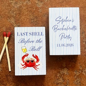 May include: Two matchboxes with blue and white striped patterns. One box reads "LAST SHELL Before the Bell" with a crab illustration. The other box says "Sophia's Bachelorette Party 11.06.2026". Two matches with red tips are next to the matchboxes.