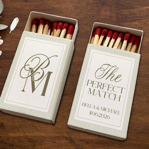 May include: Two matchboxes with red-tipped matches. One box has the initials "BM" in green script, the other reads "The PERFECT MATCH" with a date. The boxes are white with green striped sides and sit on a dark wood surface.