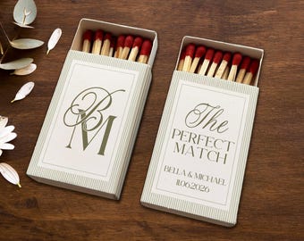 May include: Two matchboxes with red-tipped matches. One box has the initials "BM" in green script, the other reads "The PERFECT MATCH" with a date. The boxes are white with green striped sides and sit on a dark wood surface.