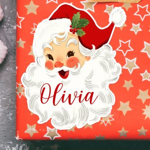 May include: A festive Christmas gift with a Santa Claus sticker. The sticker features a smiling Santa with a red hat, white beard, and the name "Olivia" in red script. The gift wrap is red with gold stars.