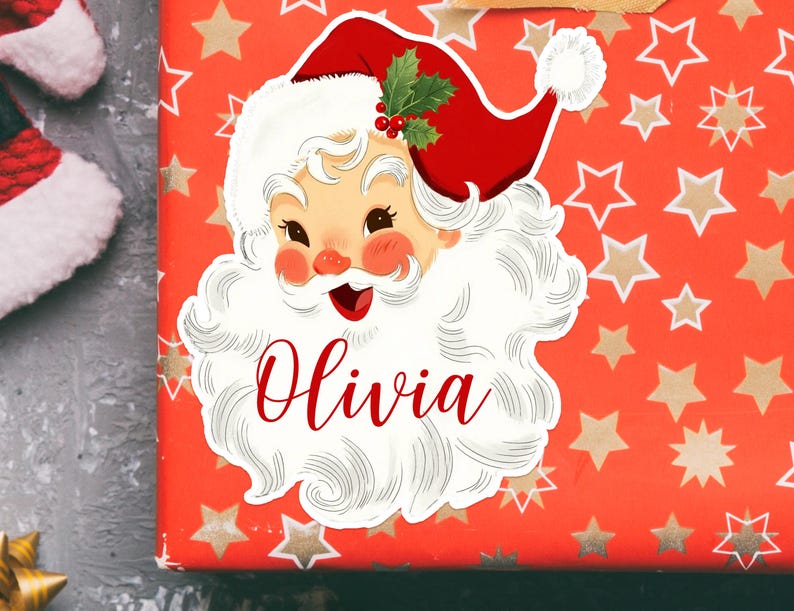May include: A festive Christmas gift with a Santa Claus sticker. The sticker features a smiling Santa with a red hat, white beard, and the name "Olivia" in red script. The gift wrap is red with gold stars.