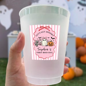 May include: A frosted plastic cup featuring a rectangular label with pink and white vertical stripes. The label displays a pink scalloped border, a ghost, a pumpkin, a cat, and the text: Let's Get Spooky, Sophia's First Boo-Day.