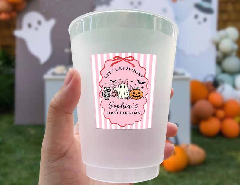 May include: A frosted plastic cup featuring a rectangular label with pink and white vertical stripes. The label displays a pink scalloped border, a ghost, a pumpkin, a cat, and the text: Let's Get Spooky, Sophia's First Boo-Day.