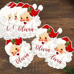 May include: A collection of Santa Claus-shaped name tags. Each tag features a smiling Santa face with a red hat and white beard. The name "Olivia" is written in red script on each tag. The tags are on a wooden surface.