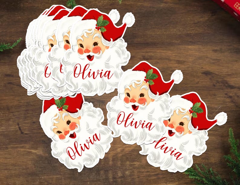 May include: A collection of Santa Claus-shaped name tags. Each tag features a smiling Santa face with a red hat and white beard. The name "Olivia" is written in red script on each tag. The tags are on a wooden surface.