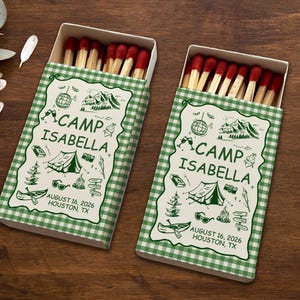 May include: Two matchboxes with red-tipped matches. The boxes have a green and white checkered pattern and feature the text "CAMP ISABELLA" with camping-themed illustrations. The date "AUGUST 16, 2026" and location "HOUSTON, TX" are also printed on the boxes.