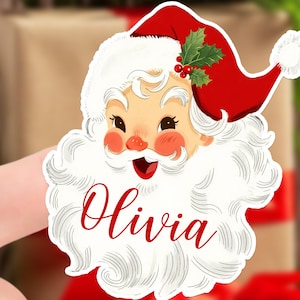 May include: A Santa Claus-themed sticker with a red hat, white beard, and the name "Olivia" in red script. The sticker features a smiling Santa face with rosy cheeks and a sprig of holly on his hat, perfect for Christmas decorations.