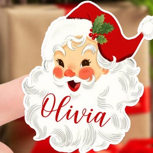 May include: A Santa Claus-themed sticker with a red hat, white beard, and the name "Olivia" in red script. The sticker features a smiling Santa face with rosy cheeks and a sprig of holly on his hat, perfect for Christmas decorations.
