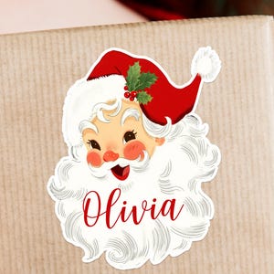 May include: A sticker featuring a vintage Santa Claus design with a red hat, white beard, and the name "Olivia" in red script. The sticker is placed on a wrapped gift with a red ribbon.