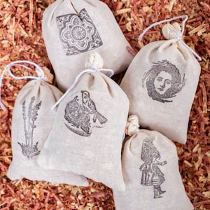 May include: Five off-white fabric drawstring pouches featuring black ink illustrations. The designs include a geometric tile pattern, a bird in a nest, a sprig of lavender, a human face surrounded by swirling lines, and a standing figure in a dress.